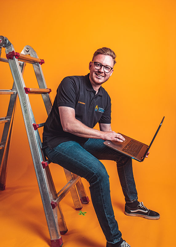 Ein junger Mann sitzt auf einer Leiter, trägt ein Laptop, ein schwarzes Polo mit Namensstick „S. Schiller“ vor orangefarbenem Hintergrund.
